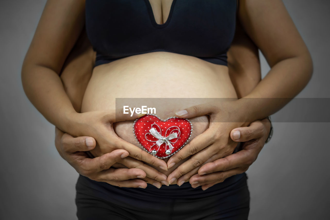 Cropped hands of man with pregnant girlfriend holding red heart shape on abdomen against gray background