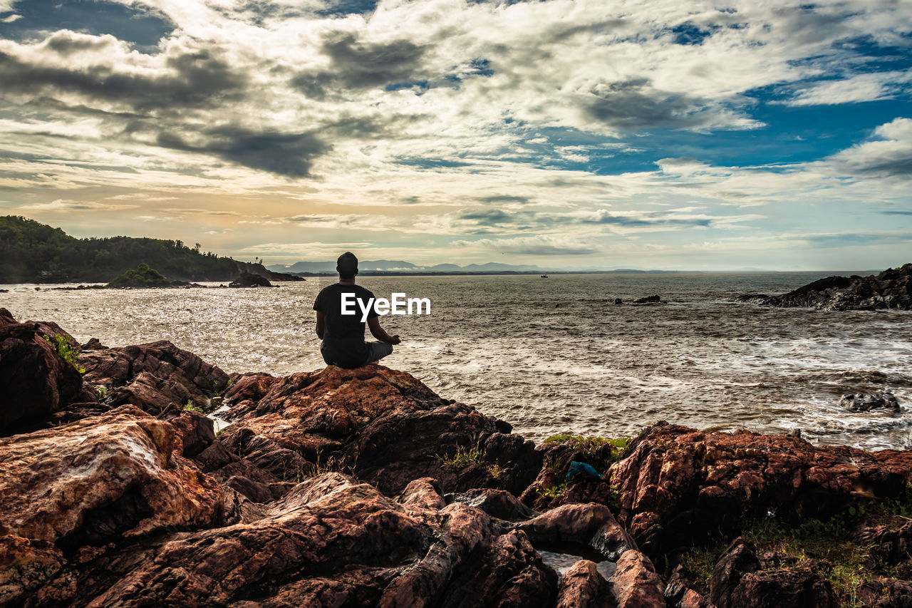 Rear view of man sitting on rock by sea against sky