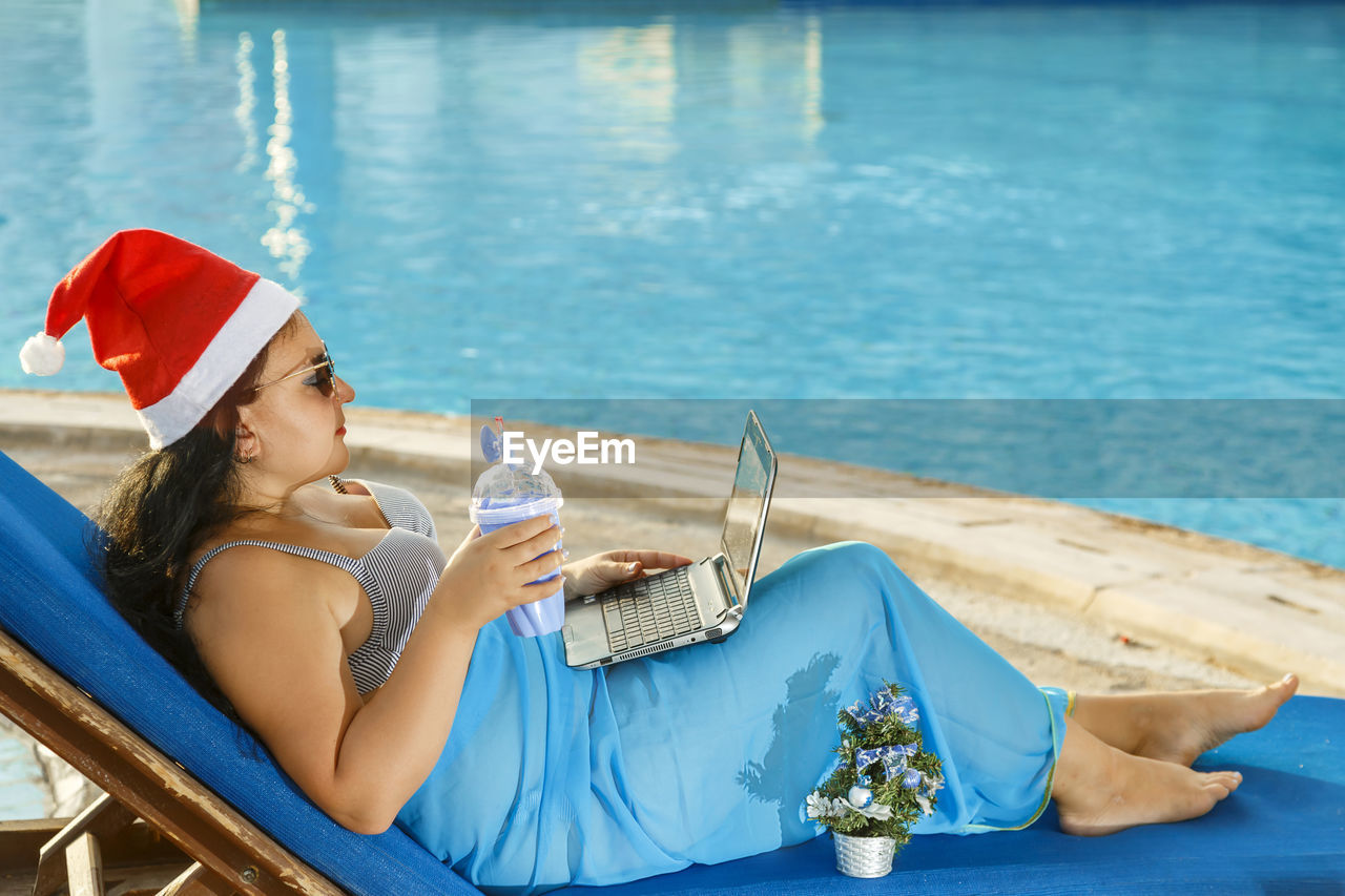 Side view of woman wearing santa hat using laptop by swimming pool