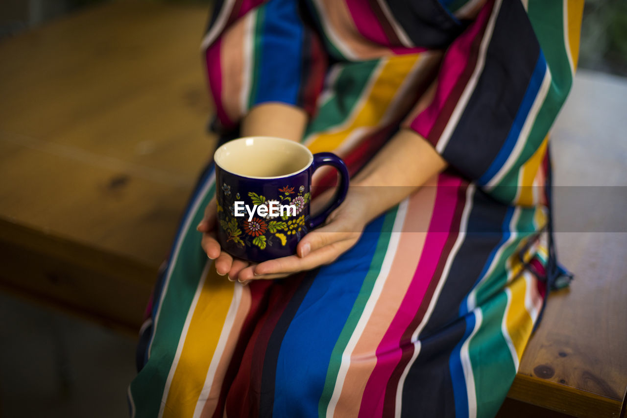 Woman in multi colored clothing holding coffee cup while sitting on table at home