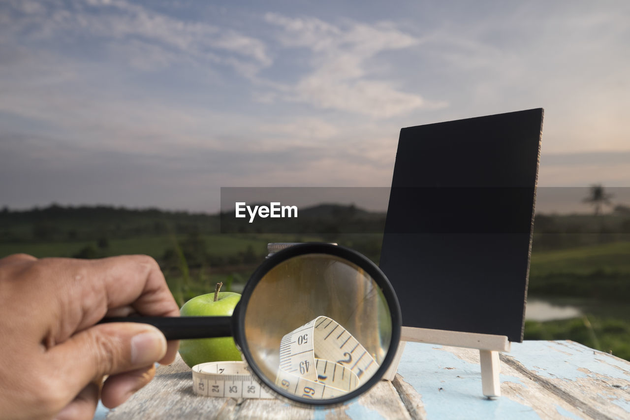 Cropped hand holding magnifying glass at table against sky