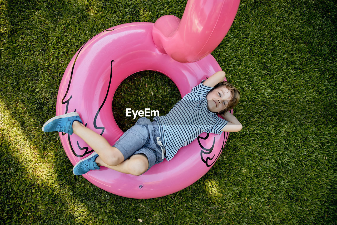 Blond boy in blue clothes, lying on pink inflatable flamingo on grass.