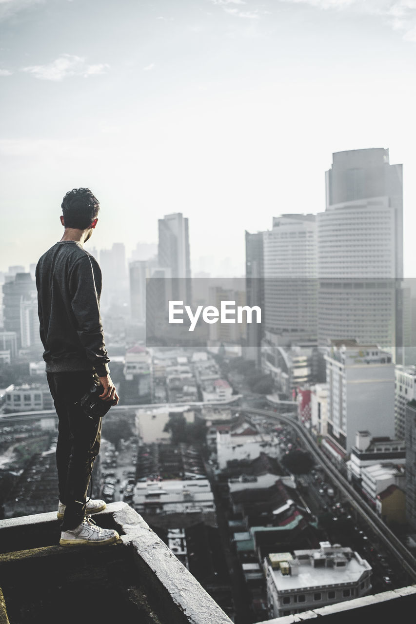 Full length of man standing on building terrace by cityscape against sky