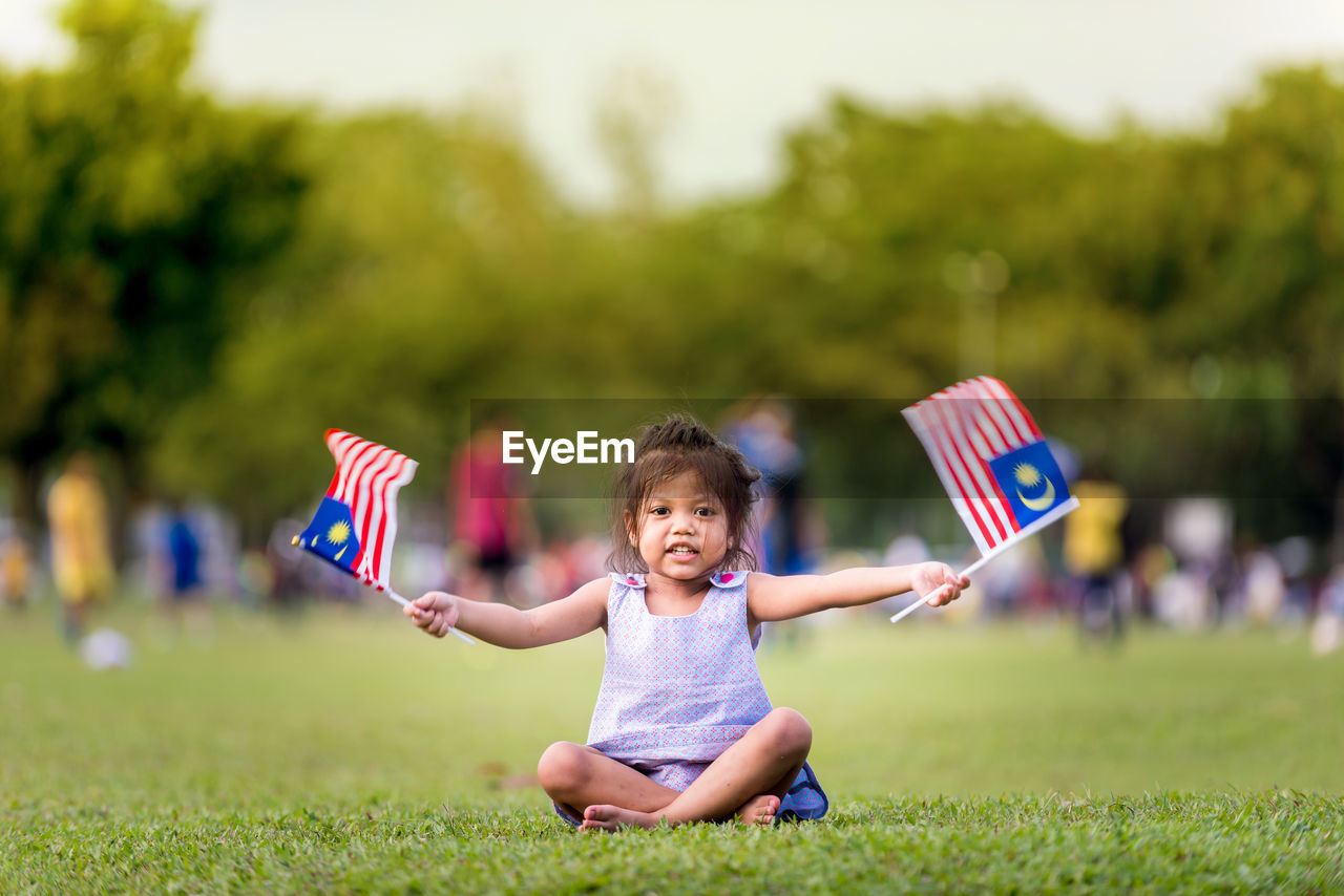 Portrait of cute girl sitting with malaysian flags on grass at park