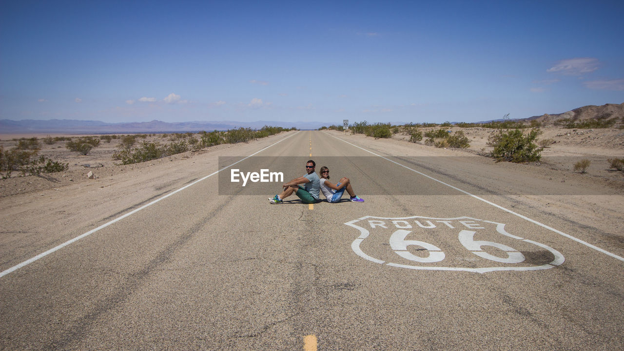 Side view of couple sitting on road against blue sky