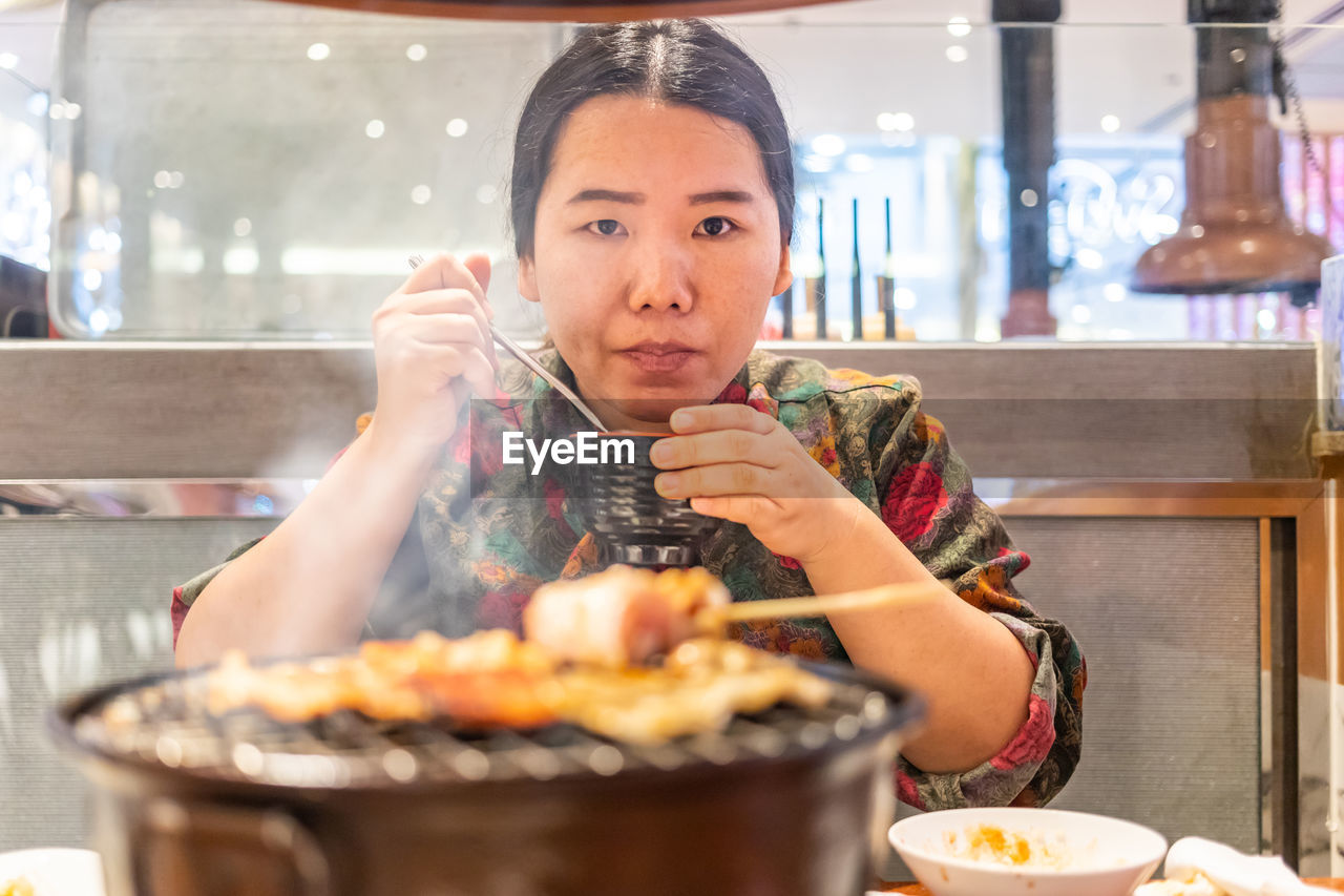 Portrait of woman eating food by barbecue grill in restaurant
