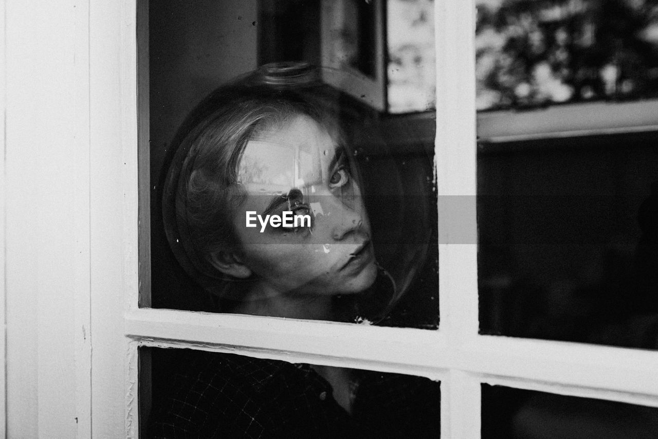 PORTRAIT OF YOUNG WOMAN LOOKING THROUGH WINDOW AT HOME