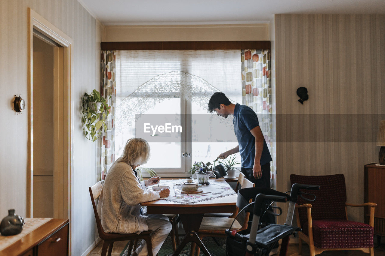 Male healthcare worker watering plants while senior woman eating food at home