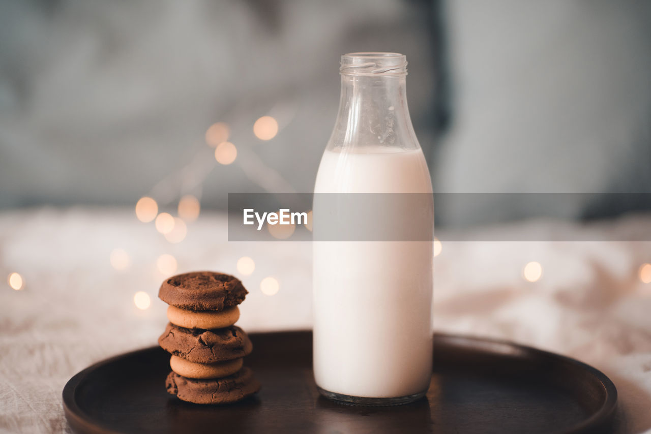 Glass bottle with fresh milk and stack of chocolate cookies on wooden tray in bed closeup. 