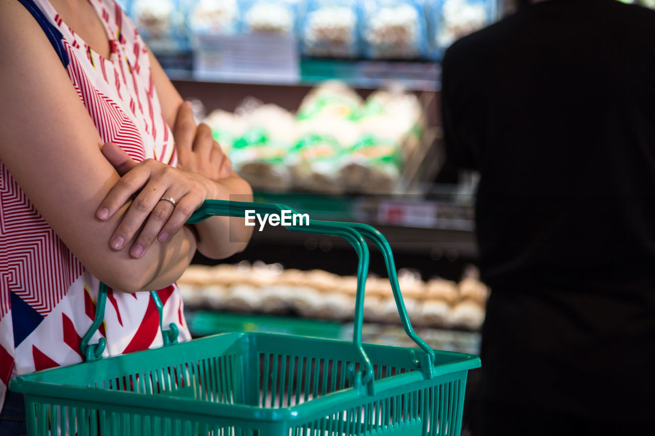 Midsection of woman holding shopping basket with man in background at supermarket