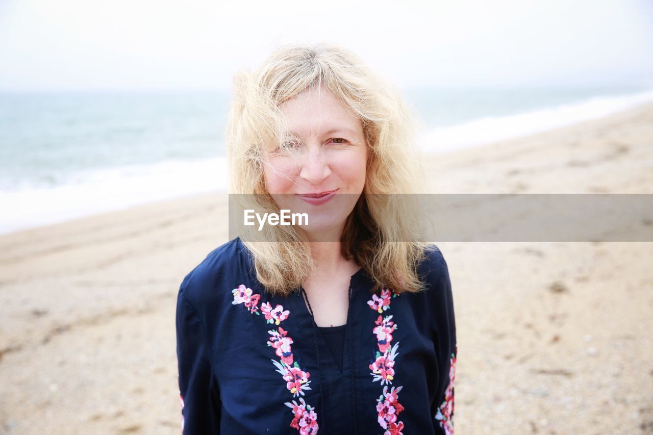 Portrait of smiling mature woman standing at beach against sky