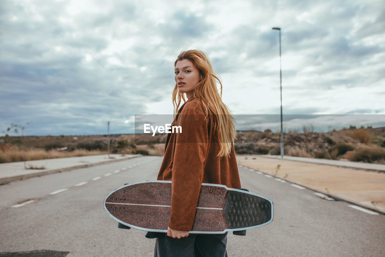 Side view of young female skater with long blond hair in trendy outfit standing on asphalt road with cruiser skateboard in hand against cloudy sky in countryside