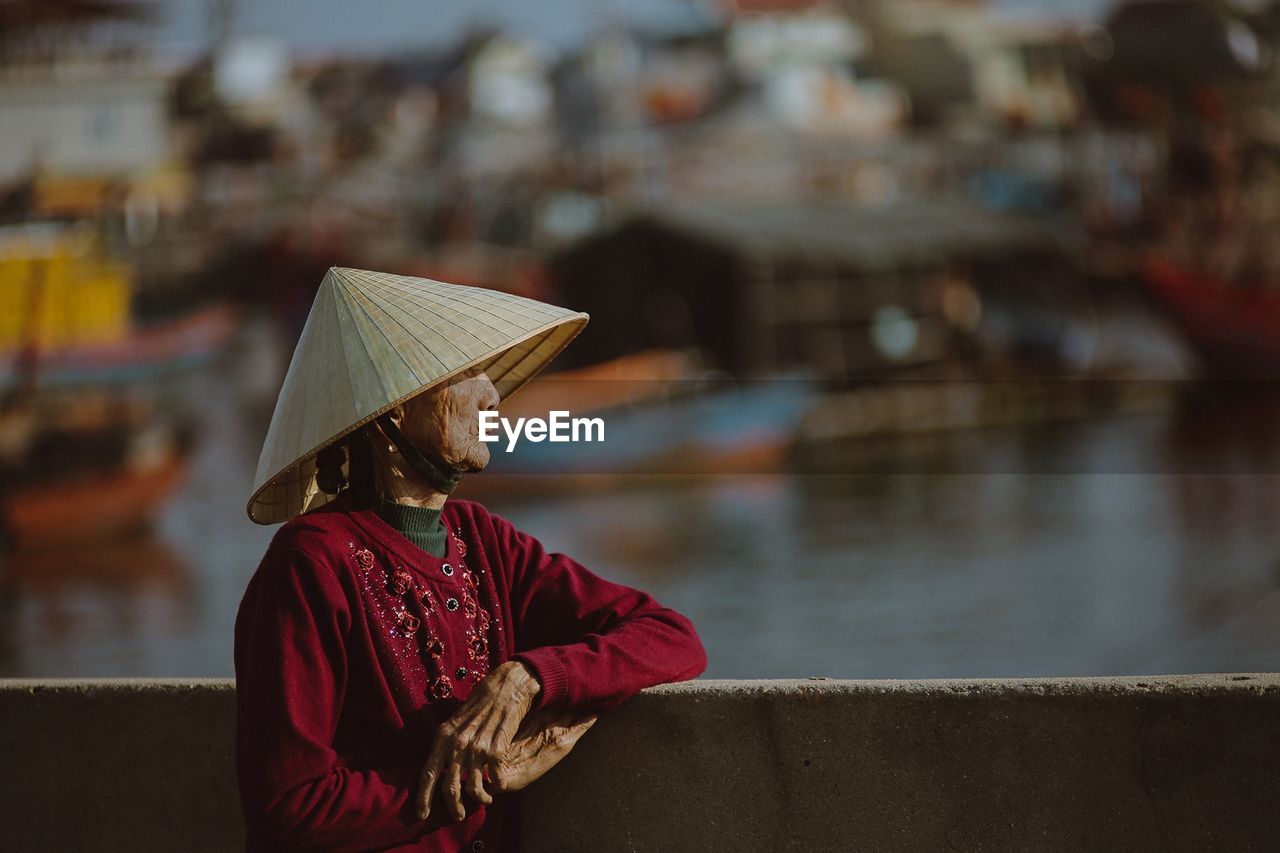 one person, water, adult, clothing, hat, women, architecture, nautical vessel, focus on foreground, waist up, nature, traditional clothing, city, day, transportation, outdoors, lifestyles, person, occupation, rear view, red, temple, holding, rain, asian style conical hat, river, standing