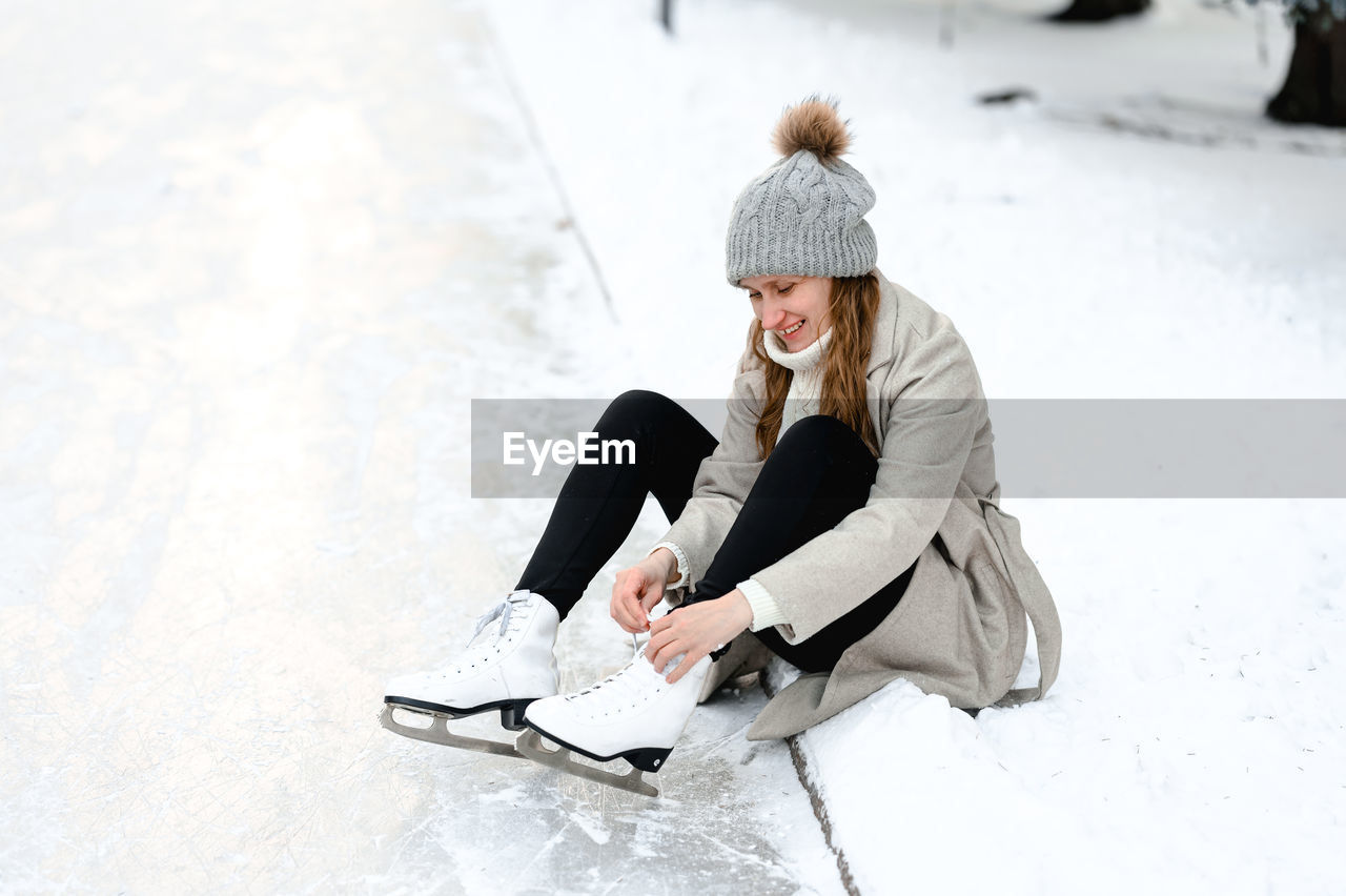 Woman in a coat came to skate on an ice rink, she sits in the snow and ties up her ice skates