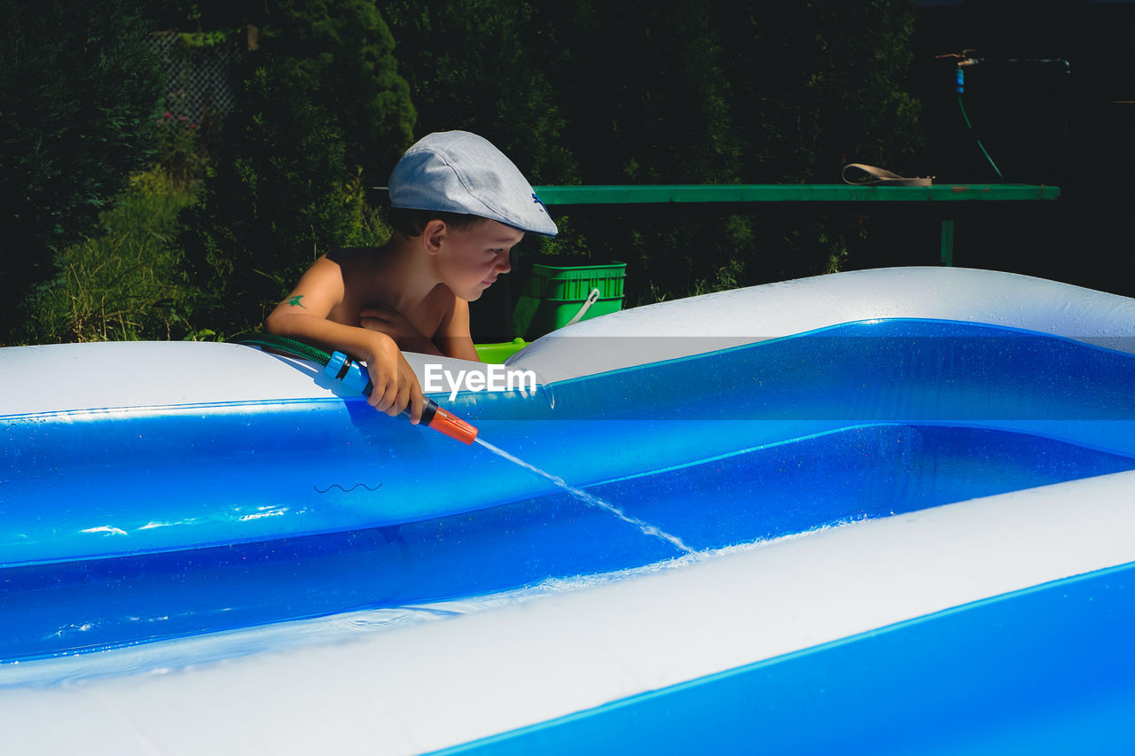 Boy spraying water in wading pool with garden hose during summer