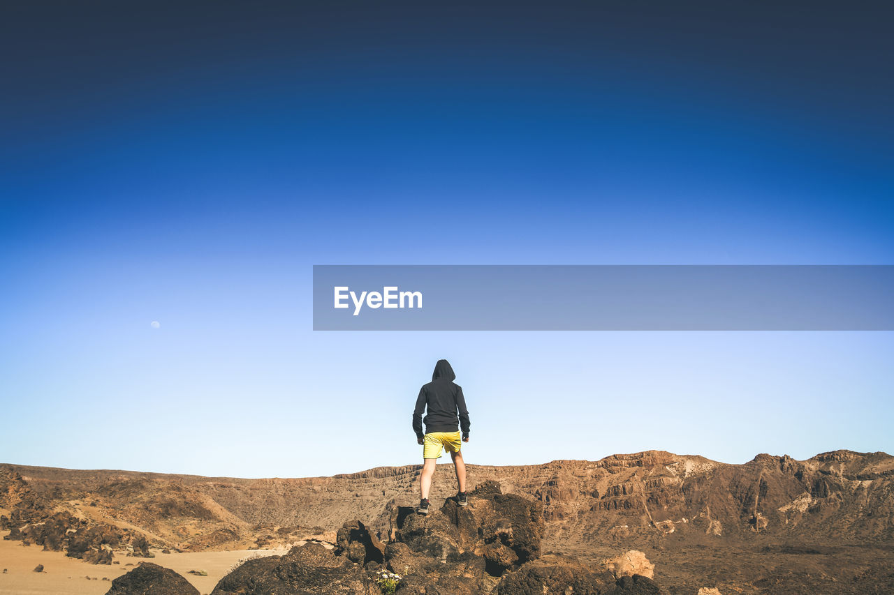 Rear view of boy standing on rocks against clear sky