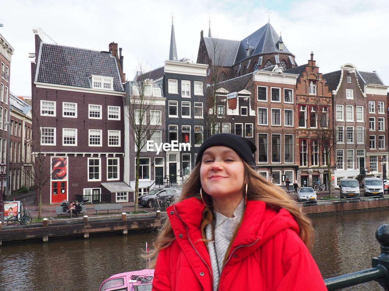 PORTRAIT OF SMILING YOUNG WOMAN WITH BUILDINGS IN CANAL