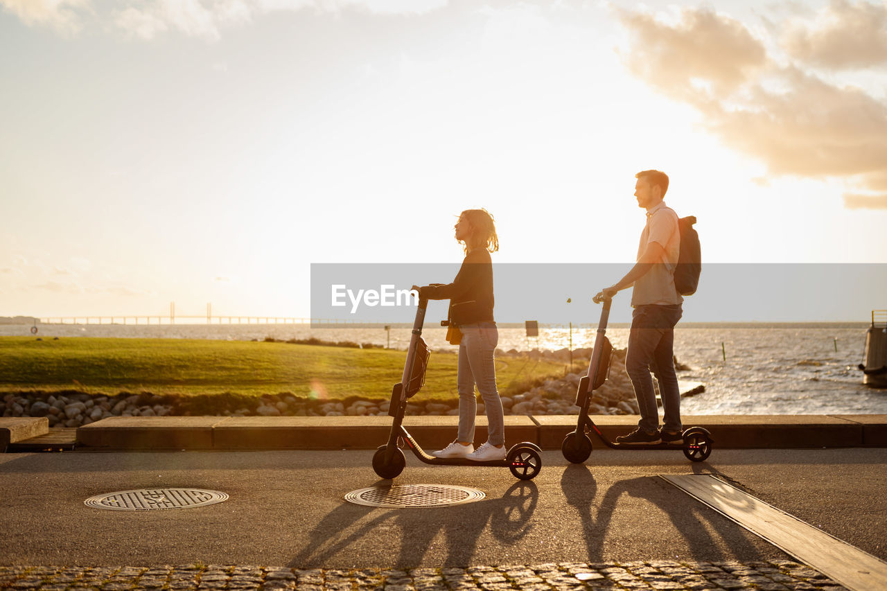 Full length of couple enjoying on electric push scooters by sea against sky during sunset