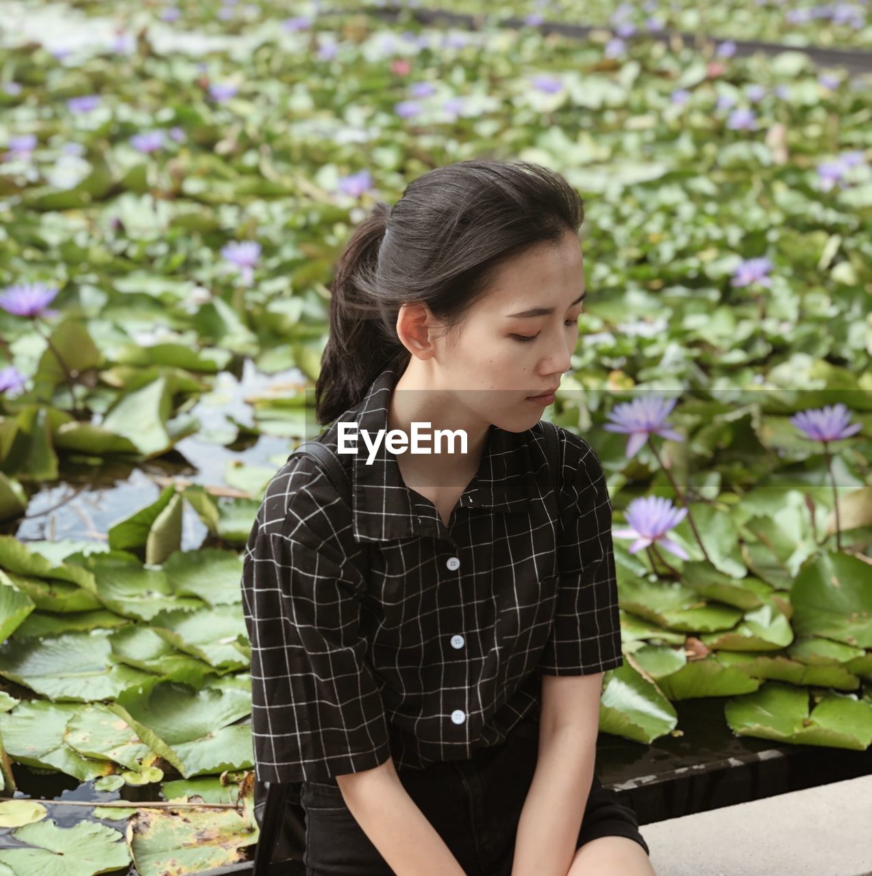 Sad young woman looking down while sitting by lotus plants in pond