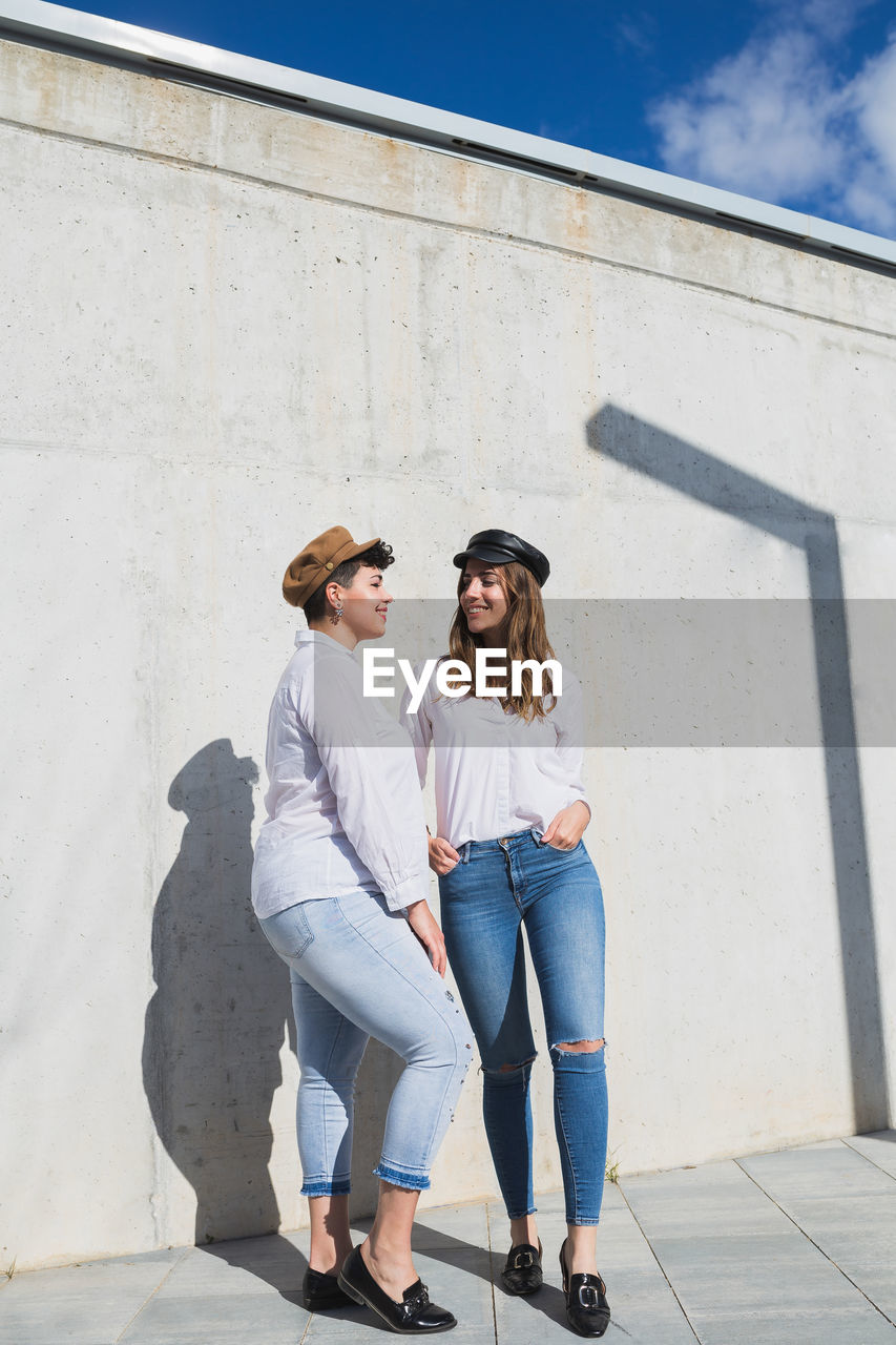 Full body of young positive female friends in trendy outfits and hats standing looking at each other on walkway near gray wall in sunny day under blue sky