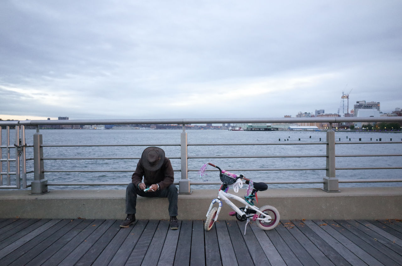 Man wearing hat sitting on pier by children bicycle while using smart phone