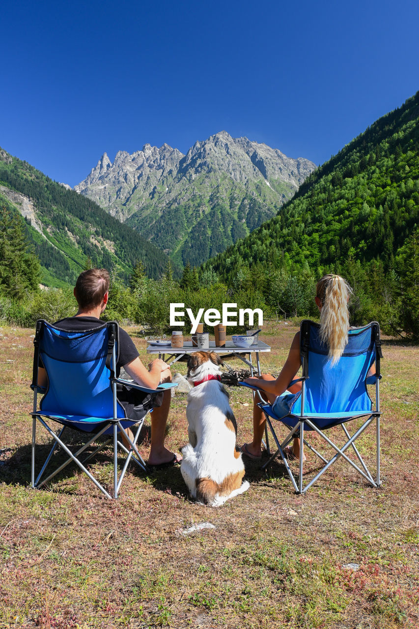 A young couple and a dog sitting in a field looking at mountains.