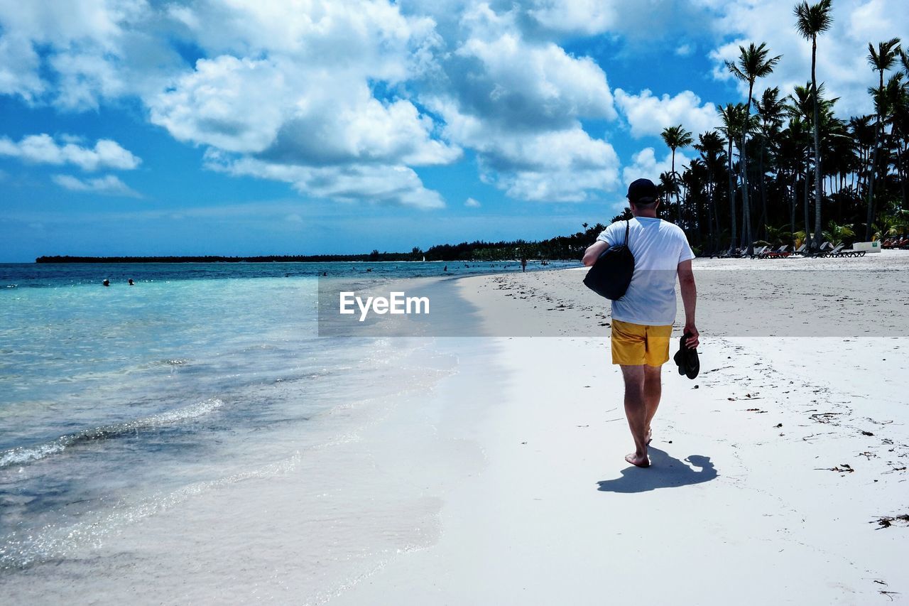 Rear view of mature man walking at beach against cloudy sky during sunny day