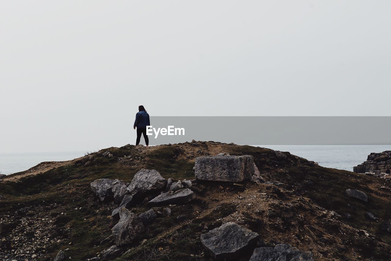 MAN STANDING ON ROCK BY SEA AGAINST SKY