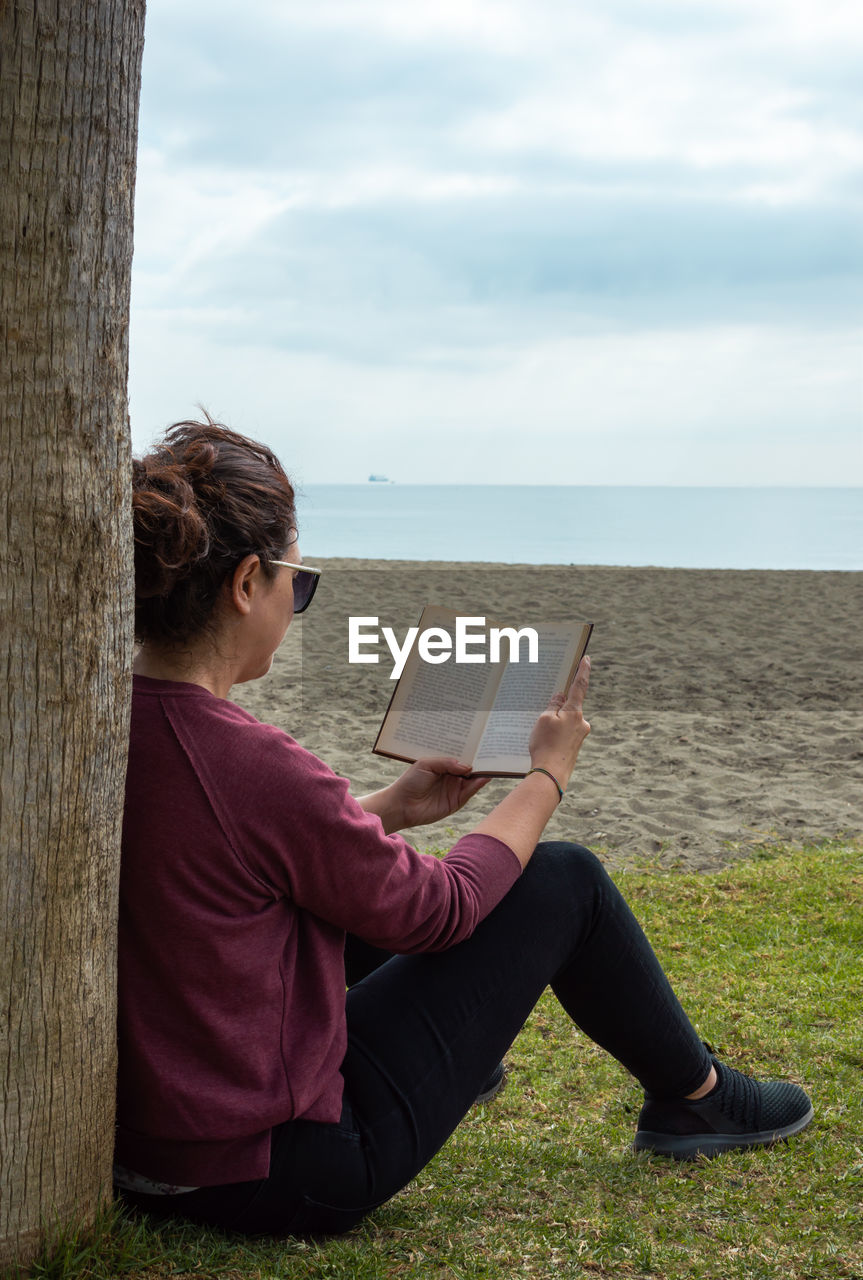 WOMAN SITTING ON BOOK AT SEA SHORE AGAINST SKY