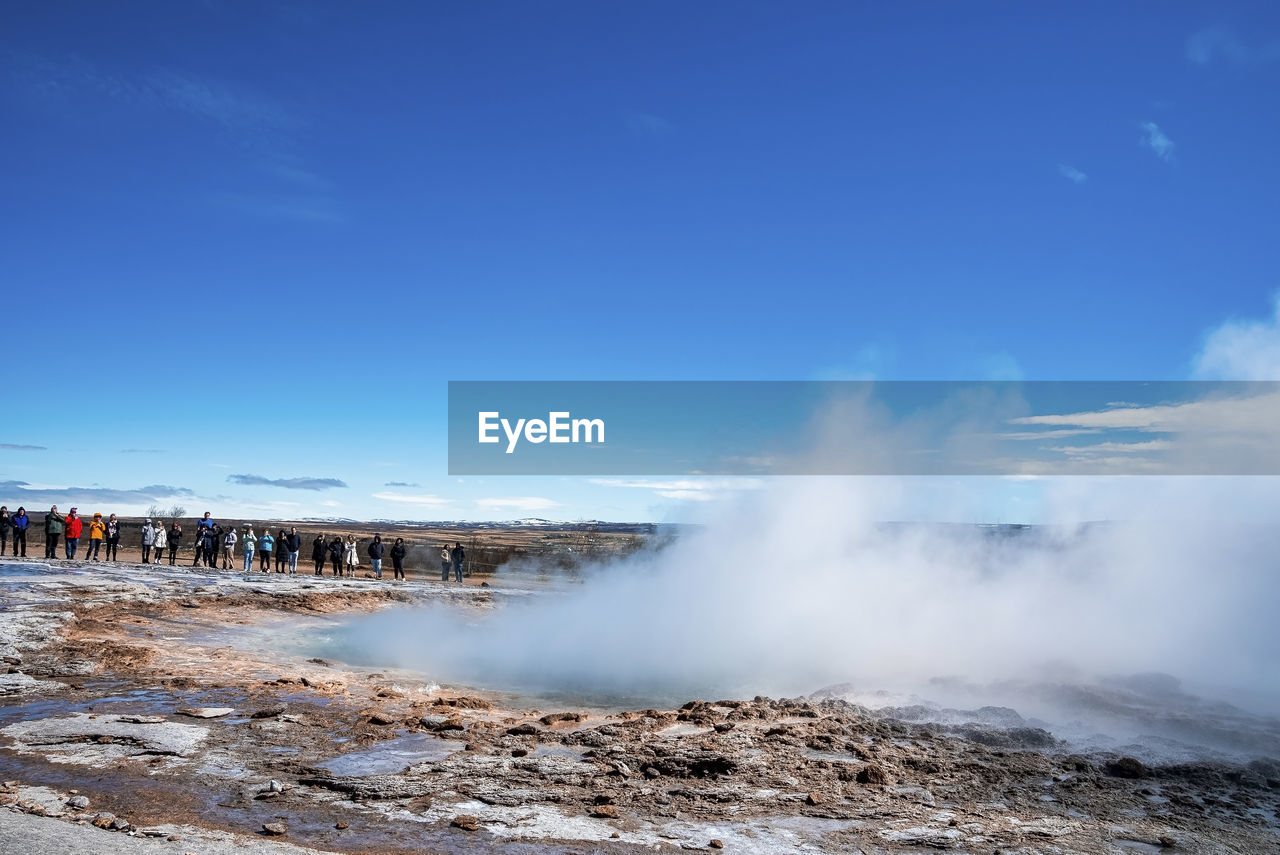 Tourists exploring strokkur geyser eruption in valley against blue sky