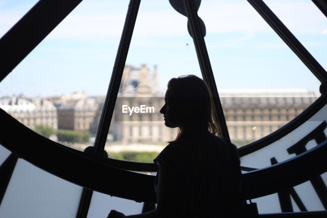 REAR VIEW OF SILHOUETTE WOMAN LOOKING AT CITYSCAPE SEEN THROUGH WINDOW