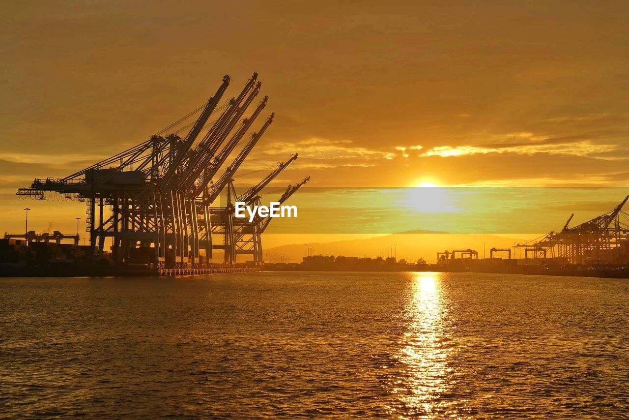 Cranes at harbor against sky during sunset