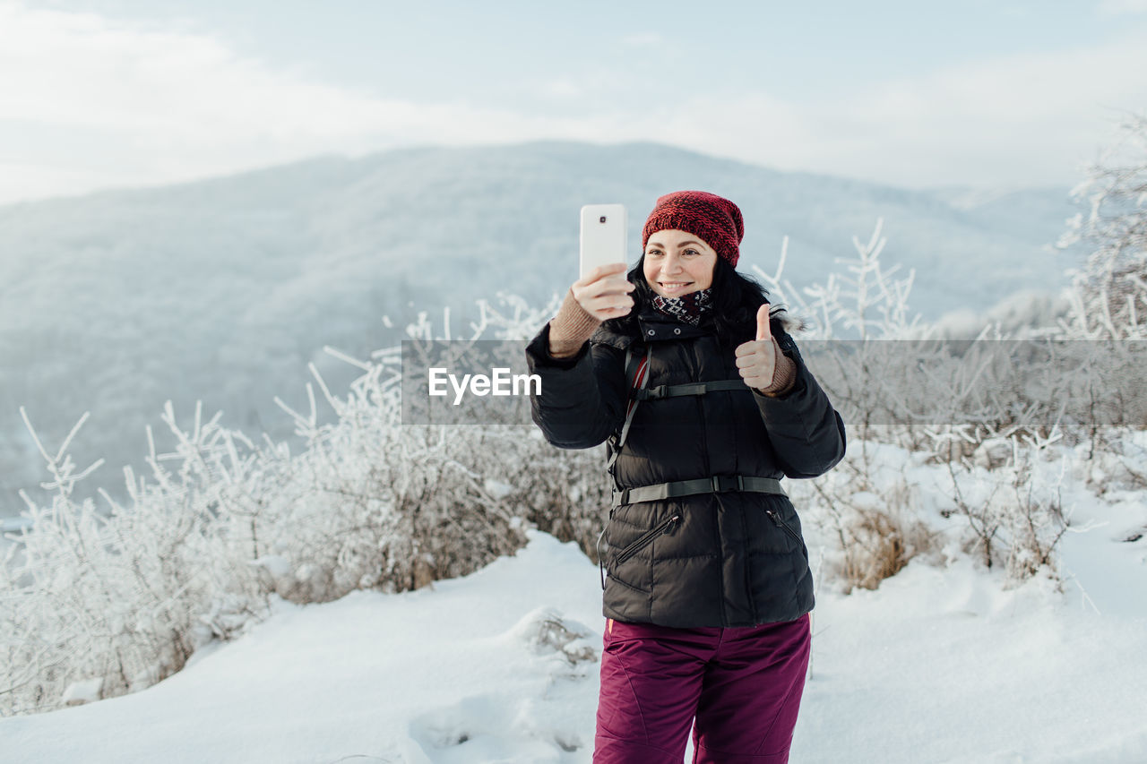 Smiling woman in warm taking selfie while standing on snowcapped mountain