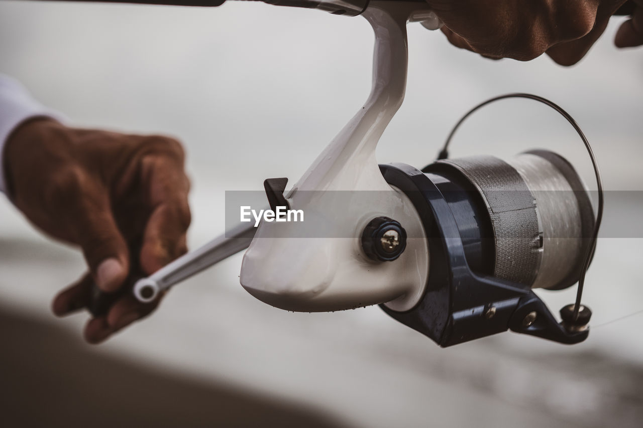 Crop anonymous man turning metal handle of special spinning reel standing on coast while fishing in nature on blurred background