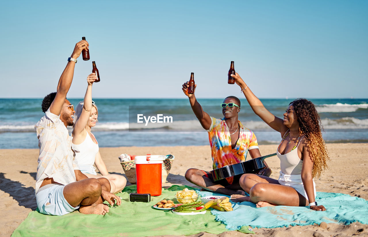 Multiracial men and women in casual clothes rising up bottles of beer and proposing toast while sitting cross legged on blanket during picnic on beach on summer weekend day