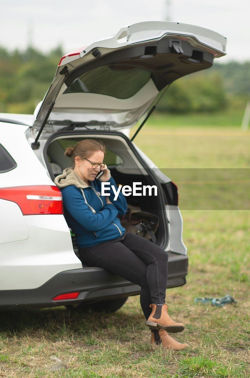 Woman talking on phone while sitting in car trunk on field