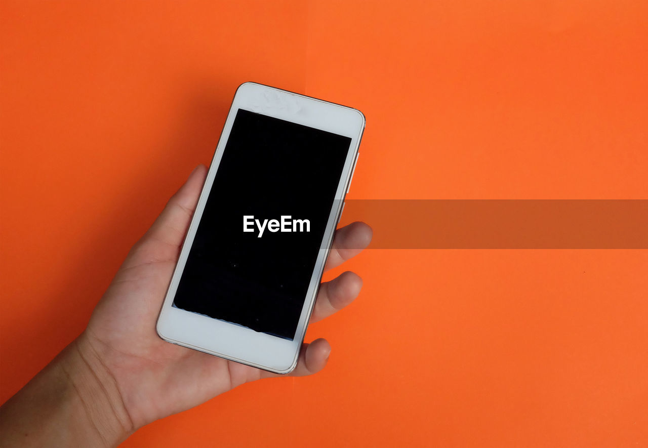 CLOSE-UP OF HAND HOLDING MOBILE PHONE OVER ORANGE BACKGROUND