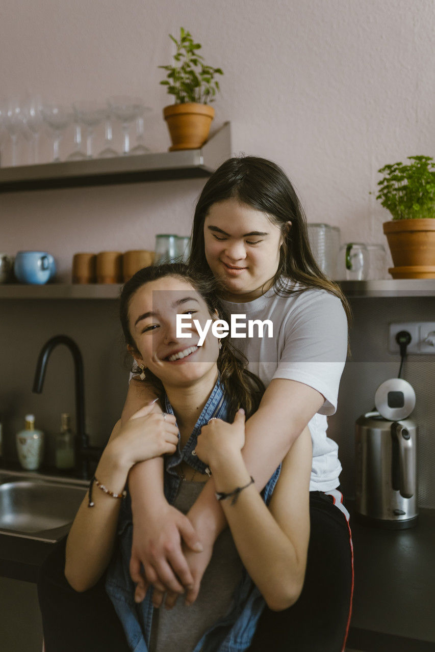 Young woman with down syndrome embracing sister from behind in kitchen at home