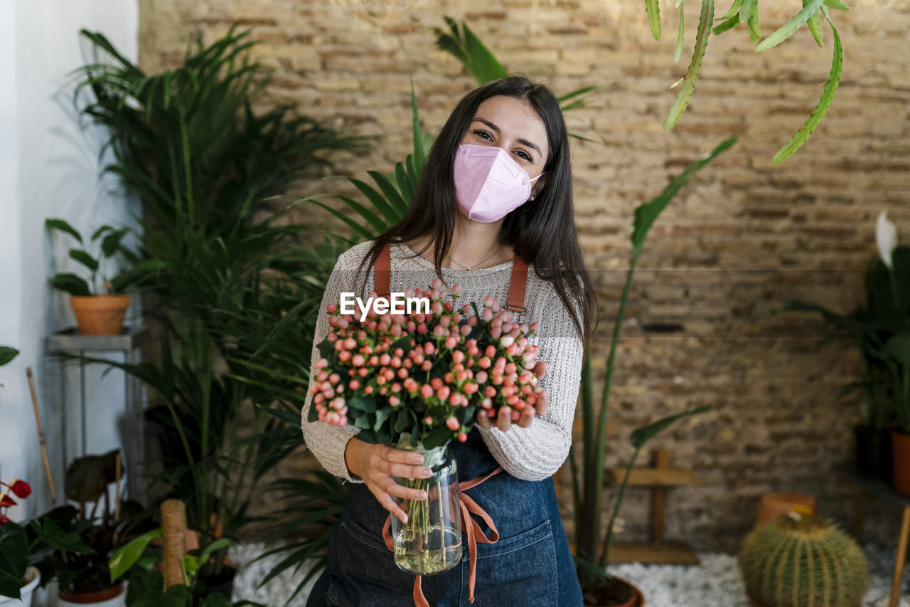Young female florist with protective face mask holding hypercium flower vase at shop