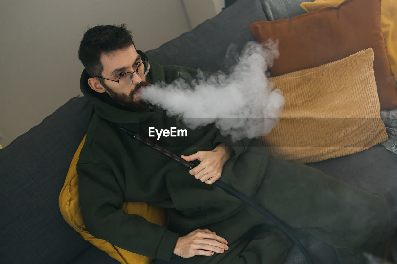 smoke, adult, one person, men, indoors, smoking, person, clothing, smoking issues, activity, cigarette, communication, glasses, beard, social issues, holding, young adult, facial hair, sitting, furniture, lifestyles, bad habit, sofa, front view, eyeglasses, waist up, mammal, leisure activity
