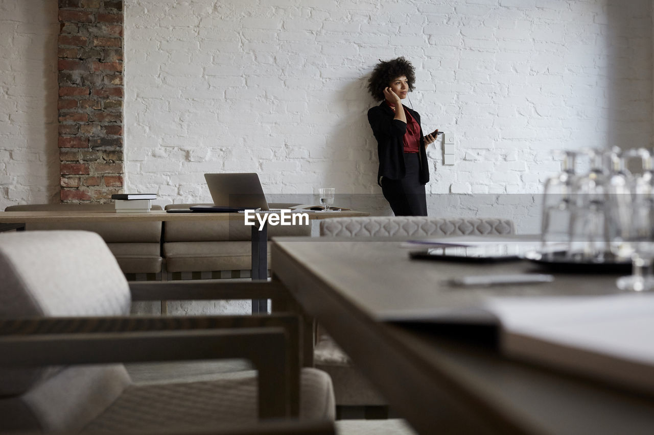 Confident young female lawyer talking on mobile phone while leaning on white wall at office