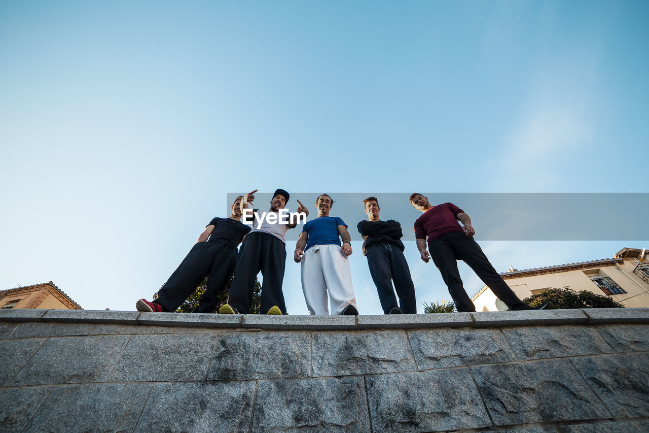 Low angle view of male friends standing on retaining wall against sky