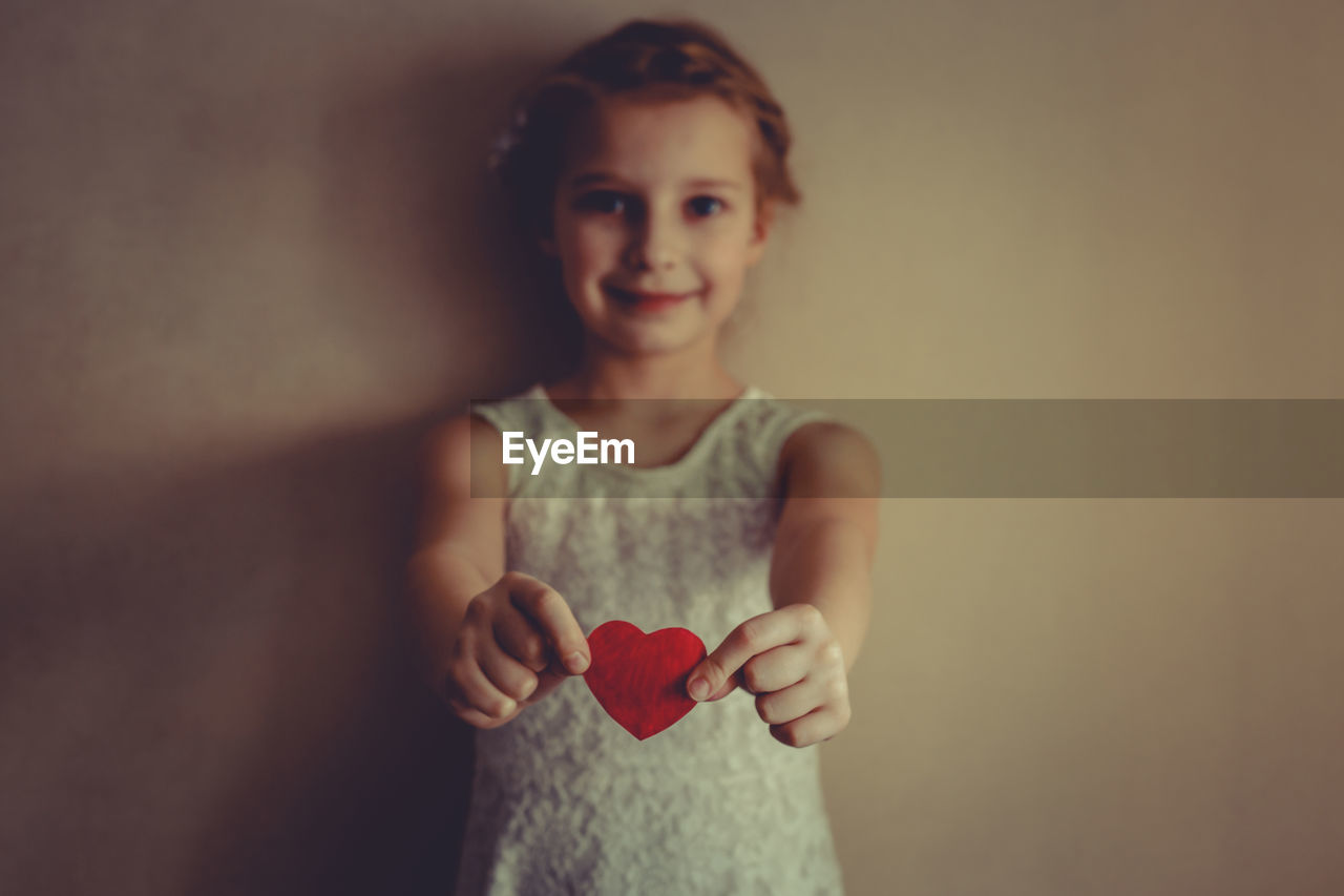 Portrait of girl holding heart shape against wall