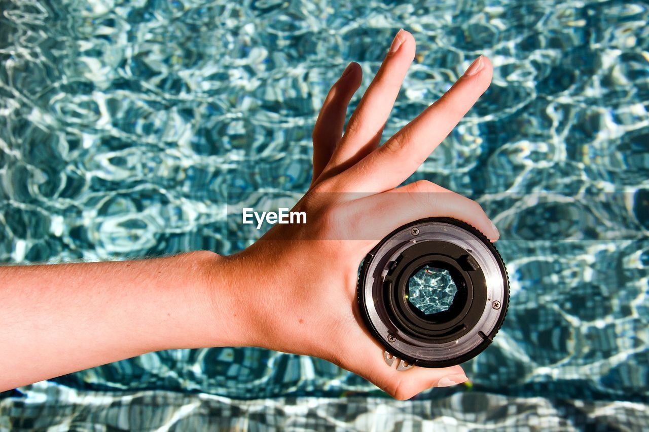 Cropped hand of person holding lens against water at swimming pool