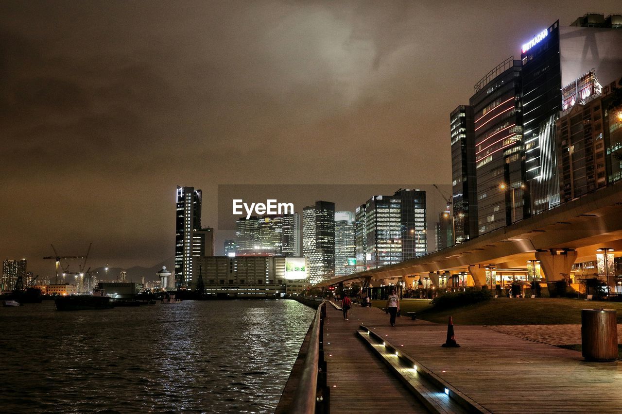 Illuminated skyscrapers and promenade by sea against sky