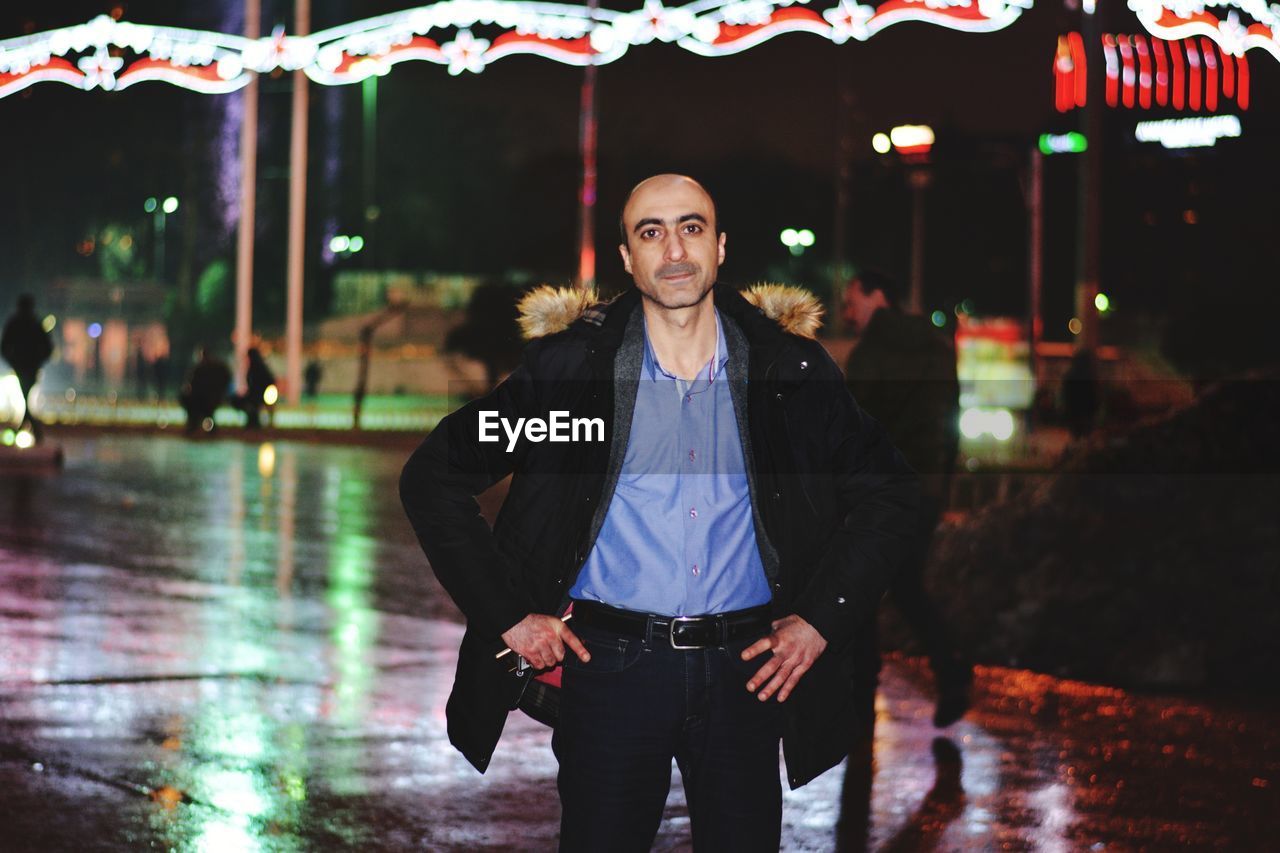 Portrait of man with hands on hip standing on footpath at night