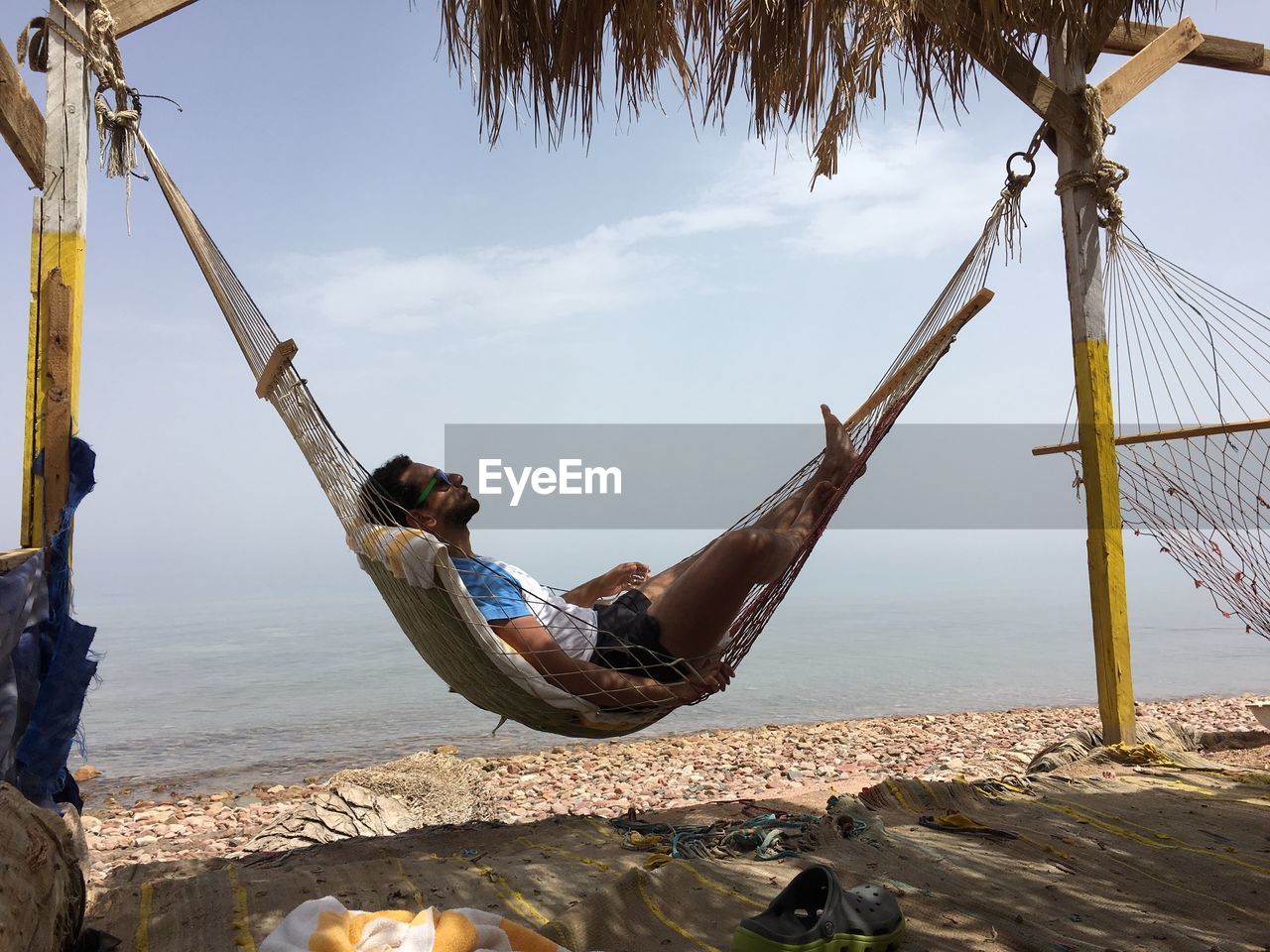 Side view of young man relaxing on hammock at beach against sky