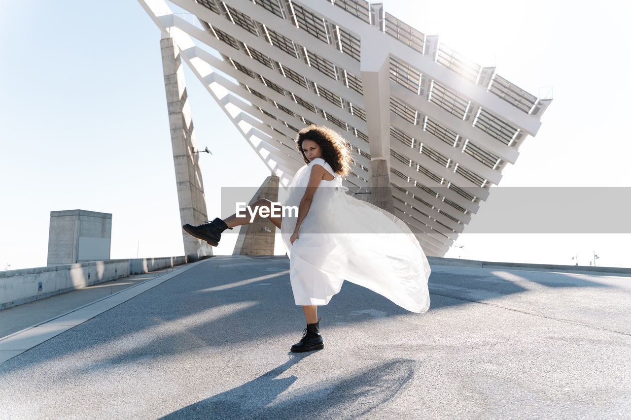 Full body of cool bride in brutal boots dancing looking at camera on paved waterfront with solar batteries on background in back