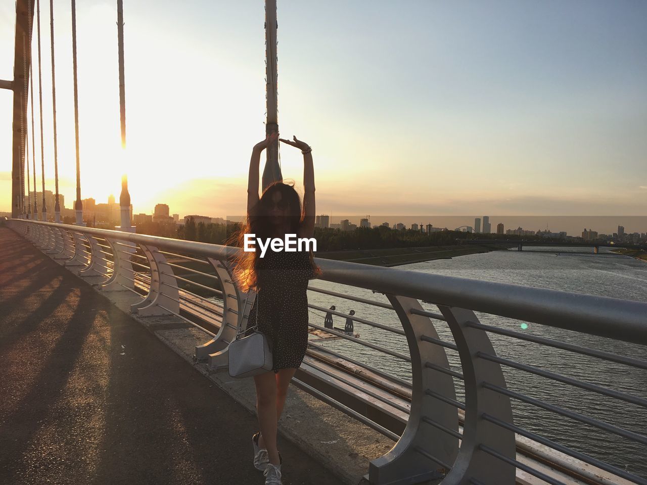 Full length of woman with arms raised at bridge over river during sunset