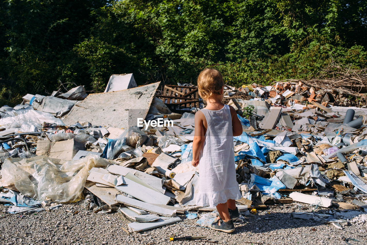 Little girl at a dump among a heap of scattered garbage in the forest.